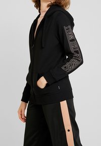 Sweat à capuche noir zippé avec une poche frontale, présentant un texte vertical blanc sur la manche gauche et une capuche doublée. Matière en mélange de coton lisse.