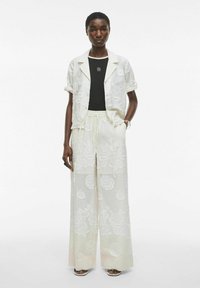 Chaqueta de camisa blanca con bordado floral sobre una blusa negra, combinada con pantalones claros de diseño fluido y cintura con cordón.