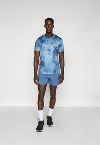 T-shirt de sport bleu avec un motif texturé, à manches courtes et un petit logo blanc, associé à un short bleu et des chaussures de course noires.