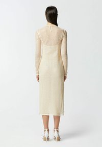 Duga, uska haljina od prozirne, teksturirane bež tkanine s dugim rukavima, ključanom pozadinskom kopčom i dužinom do sredine lista, uparena s bijelim potpeticama.