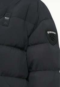 Jachetă puffer neagră cu un design matlasat, textură lucioasă și un logo brodat, rotund, negru pe umărul stâng.