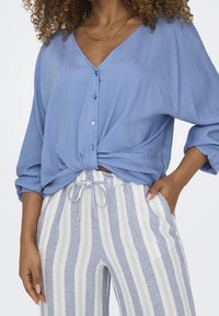 Blå knapp-up blus med V-ringning och knutdetalj vid midjan; matchad med ljusblå och vita vertikala randiga vida byxor.