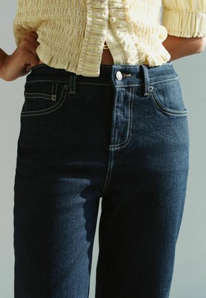 Osoba ubrana w ciemnoniebieskie dżinsy i żółty, fakturowany krótki top, z rękami opartymi na biodrach na neutralnym tle.