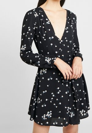 Frau trägt ein schwarzes Langarmkleid mit weißem Gänseblümchenmuster und tiefem V-Ausschnitt, die Hände vor der Taille verschränkt, gebunden mit einem passenden Stoffgürtel.