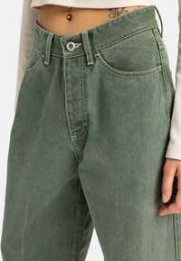 Pantalons en denim vert à taille haute, avec des coutures marquées, des poches avant et une fermeture à bouton en métal argenté. La texture est douce et légèrement brossée.