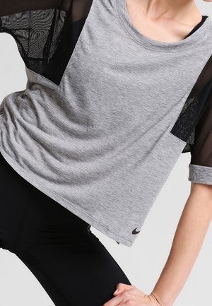 Personne portant un maillot de sport gris à manches en maille noire et des leggings noirs, une main reposant sur son genou plié.