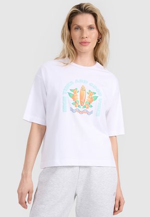 Blondynka ubrana w białą, oversize koszulkę z kolorową grafiką deski surfingowej i kwiatu oraz jasnoszare spodnie dresowe, stojąca na szarym tle.