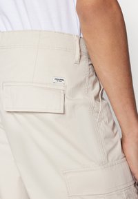 Beige cargo-housut, joissa on takatasku ja merkkilabel, sileällä pinnalla ja suoralla siluetilla. Punokset on ommeltu siististi.