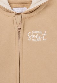Beige Zip-Hoodie mit einer cremefarbenen gefütterten Kapuze, auf der das gestickte Motiv "Du bist so süß" in Weiß auf der Vorderseite zu sehen ist. Weiches Material mit einer gemütlichen Textur.