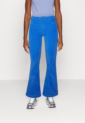 Person trägt hellblaue Schlaghosen, ein hellviolettes eng anliegendes Oberteil und weiße sowie marineblaue Sportschuhe und steht vor einem schlichten hellen Hintergrund.