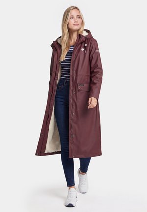 Kvinde iført en lang burgunder regnfrakke med hætte, striped skjorte, blå jeans og hvide sneakers går mod en ensfarvet baggrund.
