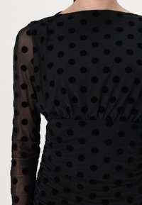 Μαύρο φόρεμα με διάφανες μακριές μανίκες και στενή μέση. Διακοσμημένο με υφή από μαύρες βούλες, με συγκεντρωμένη λεπτομέρεια στη μέση.