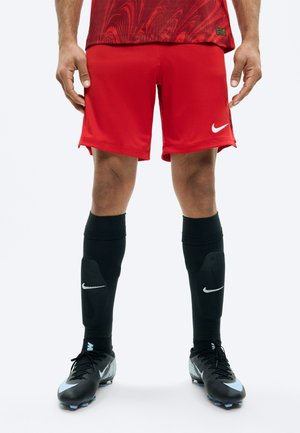 Sportowiec w czerwonych szortach sportowych, czarnych skarpetach do kolan z logo Nike oraz czarnych korkach piłkarskich z jasnoniebieskimi akcentami.