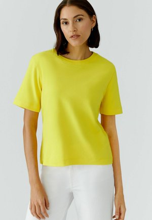 Frau mit schulterlangem dunklem Haar, die ein hellgelbes kurzärmeliges Hemd und weiße Hose trägt, steht vor einem schlichten hellen Hintergrund.