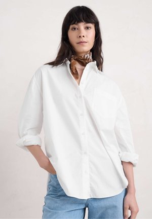 Witte button-down shirt met een borstzak, opgerolde mouwen en een gladde textuur, gecombineerd met lichtblauwe jeans en een bruine sjaal met patroon.