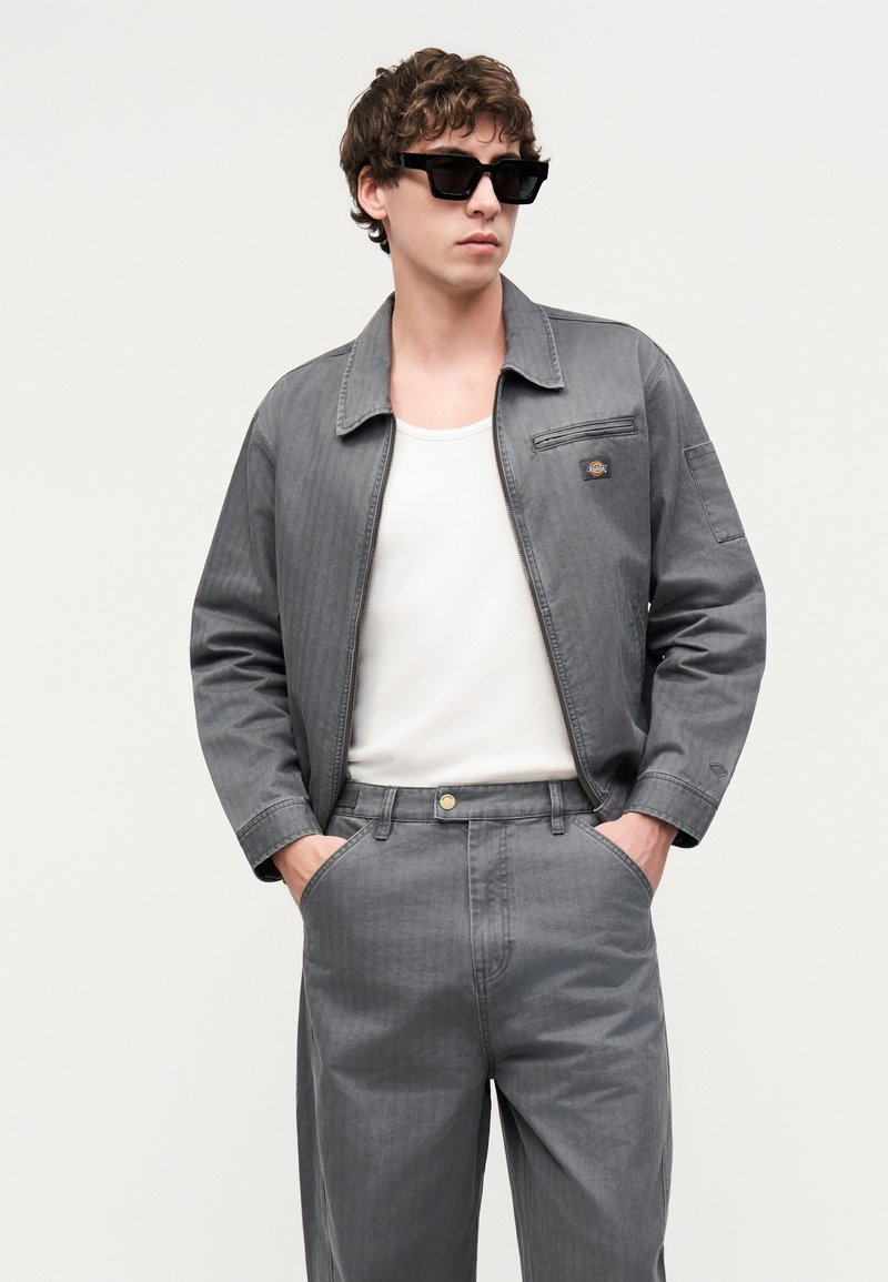 Joven con gafas de sol negras, chaqueta y pantalones vaqueros grises, camiseta blanca, de pie con las manos en los bolsillos delante de un fondo liso.