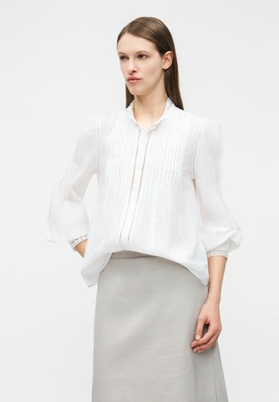 Γυναίκα που φοράει ένα λευκό μπλουζάκι με μακριά μανίκια και κάθετες δαντελένιες ρίγες, καθώς και μια ανοιχτόγρια φούστα, στέκεται με το ένα χέρι στην τσέπη.