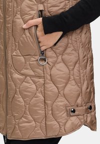 Gilet imbottito senza maniche di colore beige con tasca con zip, mano dentro e dettagli in metallo, mostra una texture liscia e un motivo arrotondato.