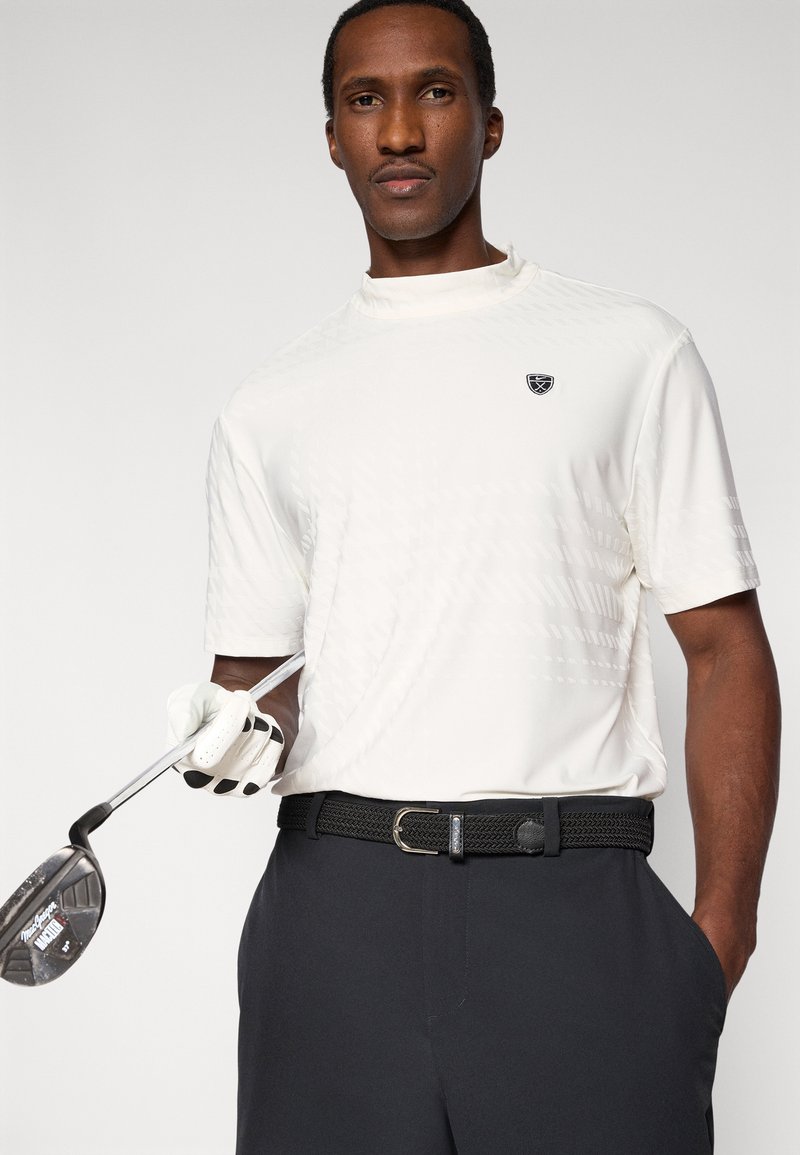 Maglietta da golf bianca leggera con motivo testurizzato, maniche corte e piccolo logo. Cintura nera intrecciata con fibbia d'argento, abbinata a pantaloni scuri.