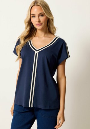 Blonde Frau trägt ein marineblaues V-Ausschnitt-Top mit weiß gestreiftem Rand und dunkelblaue Hose, steht vor hellem Hintergrund.