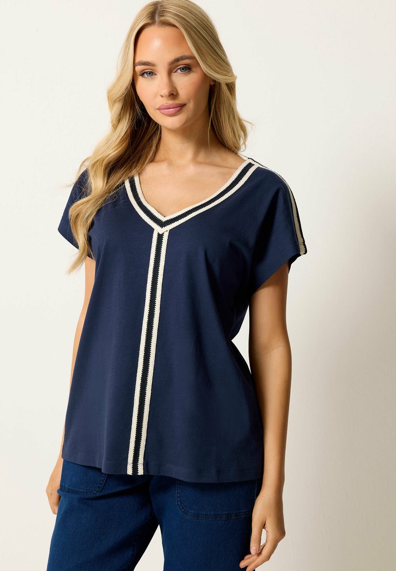 Femme blonde portant un haut bleu marine à col en V avec une bordure rayée blanche et un pantalon bleu foncé, debout devant un fond clair.