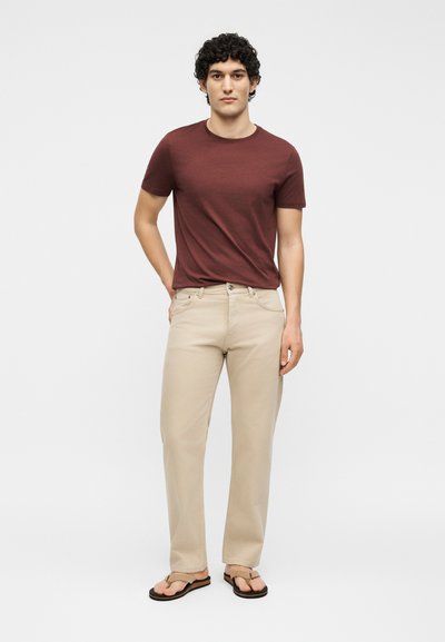 Jeune homme aux cheveux bouclés portant un t-shirt bordeaux, un pantalon beige et des sandales beiges, debout devant un fond blanc uni.