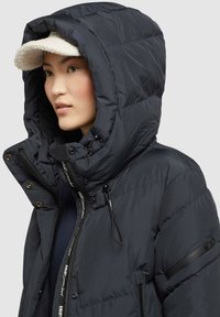 Giacca puffer blu navy con un ampio cappuccio, dotata di cerniera e bottoni a scatto. Tessuto testurizzato con cuciture a contrasto e una tasca laterale.