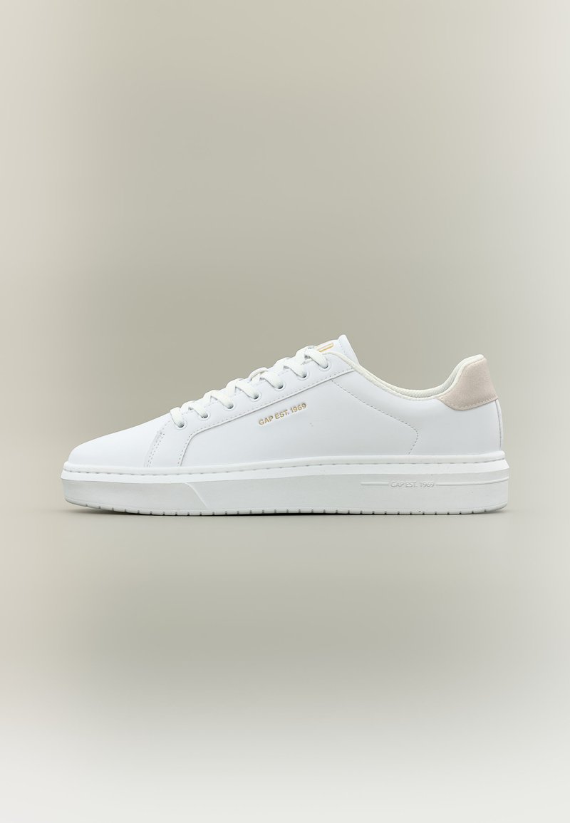 Baskets basses blanches à lacets, avec un onglet de talon beige et l'inscription dorée "GAP EST. 1969" sur le côté et la semelle, sur un fond neutre.