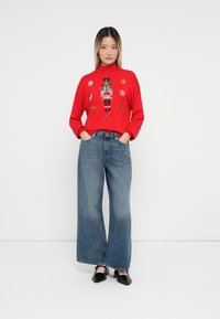 Czerwony sweter z dzianiny z haftowanym wzorem przedstawiającym dziadka do orzechów, noszony z szerokimi niebieskimi dżinsami. Czarne buty dopełniają stylizację.