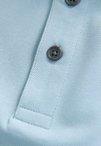 Světle modrá polo košile s texturovanou tkaninou, dvěma tmavými knoflíky a límcem. Disponuje úhledně šitým lemem a jemným vzorem.