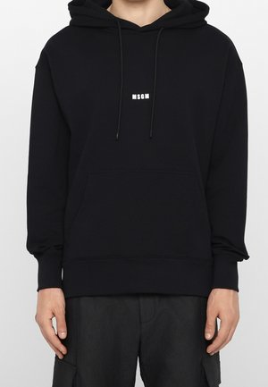 Personne portant un hoodie MSGM noir avec poche kangourou à l’avant et cordons, associé à un pantalon noir sur un fond blanc uni.