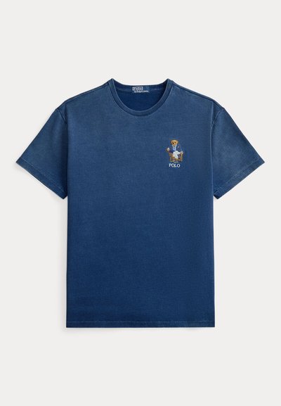 Μπλε ναυτικό κοντομάνικο T-shirt με στρογγυλή λαιμόκοψη, με ένα μικρό κεντημένο αρκουδάκι με σακάκι που κάθεται σε καρέκλα πάνω από τη λέξη «POLO» στο στήθος.