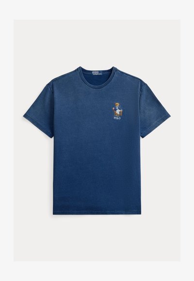 Μπλε ναυτικό κοντομάνικο T-shirt με στρογγυλή λαιμόκοψη, με ένα μικρό κεντημένο αρκουδάκι με σακάκι που κάθεται σε καρέκλα πάνω από τη λέξη «POLO» στο στήθος.