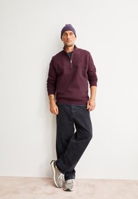 Bordeaux zip-nekt sweatshirt, donkere jeans en grijze sneakers. Het model staat tegen een witte muur en draagt een paarse beanie. Casual ontwerp.