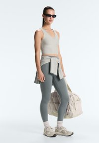 Kobieta w szarym krótkim topie, niebieskich legginsach, butach sportowych i okularach przeciwsłonecznych, z szarą bluzą przewiązaną w pasie, trzyma beżową torbę sportową.