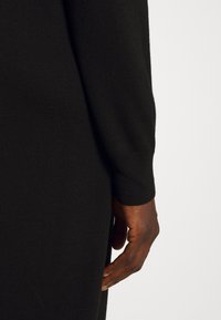 Gros plan d'une main à la peau foncée posée à côté d'un vêtement en maille noire à manches longues sur un fond clair.