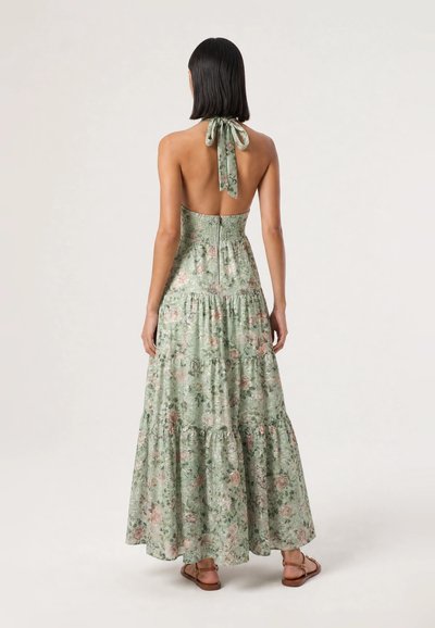 Woman wearing a backless, halter-neck, long green floral dress with tiered skirt and brown sandals, standing against a plain background.