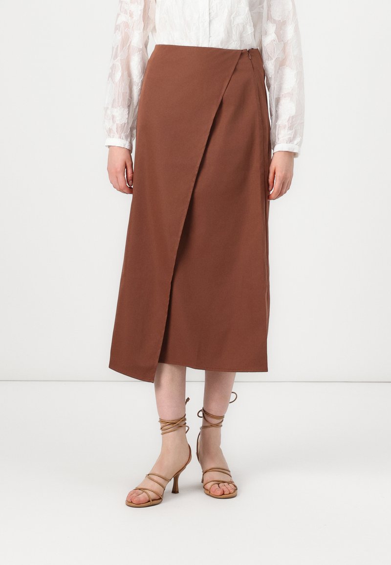 Brun omslagsskjørt laget av glatt stoff, med glidelås på siden og lengde til midten av leggen. Kombinert med beige sandaler med høye hæler og stropper.