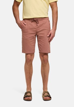 Hombre vistiendo pantalones cortos con cordón color rosa polvo, camiseta amarilla de manga corta y sandalias marrones de dos tiras, de pie con una mano en el bolsillo.