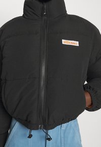 Doudoune noire avec fermeture éclair frontale et logo ellesse sur la poitrine, portée avec un pantalon bleu clair, mains partiellement dans les poches.