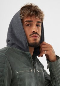 Giacca di pelle verde con fodera in felpa grigia, tasche frontali con zip e dettagli trapuntati sulle spalle. Il modello tiene una mano vicino al volto, indossando un anello.