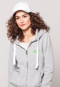 Grijze zip-up hoodie met witte koordjes, voorkeurszakken en groen logo. Draagt een plain witte T-shirt en een witte pet.