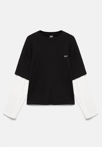 T-shirt noir à manches courtes superposé sur une chemise blanche à manches longues avec un petit logo blanc sur la poitrine, présenté sur un fond uni.