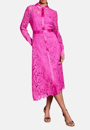 Donna che indossa un vestito midi in pizzo floreale rosa acceso con colletto in raso, polsini, patta frontale, cintura e tacchi aperti dorati su sfondo bianco.
