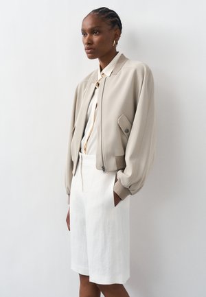 Žena s copánky má na sobě béžovou bomber bundu, bílé tričko a bílé kraťasy po kolena, stojí před jednobarevným bílým pozadím.