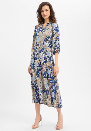 Femme portant une robe midi fleurie bleue, beige et blanche avec des manches 3/4 et des sandales à talons hauts beiges, debout devant un fond blanc.