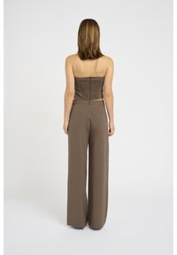 Donna in piedi con la schiena rivolta verso la macchina fotografica, vestita con un top marrone senza spalline e pantaloni ampi a vita alta marroni, su sfondo bianco.
