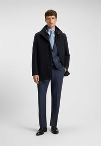 Mann in einem marineblauen Anzug mit hellblauem Hemd, gemustertem Krawatte, schwarzer Knopfjacke und schwarzen Dressschuhen, der vor einem einfarbigen, hellen Hintergrund steht.