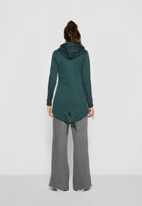 Túnica con capucha de mangas largas en verde oscuro, con un dobladillo asimétrico y un lazo ajustable en la parte posterior. Combinada con pantalones de pierna ancha grises.