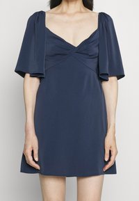 Granatowa sukienka z głębokim dekoltem w serek, krótkimi bufkami i marszczonym przodem. Wykonana z miękkiego materiału o gładkiej fakturze.